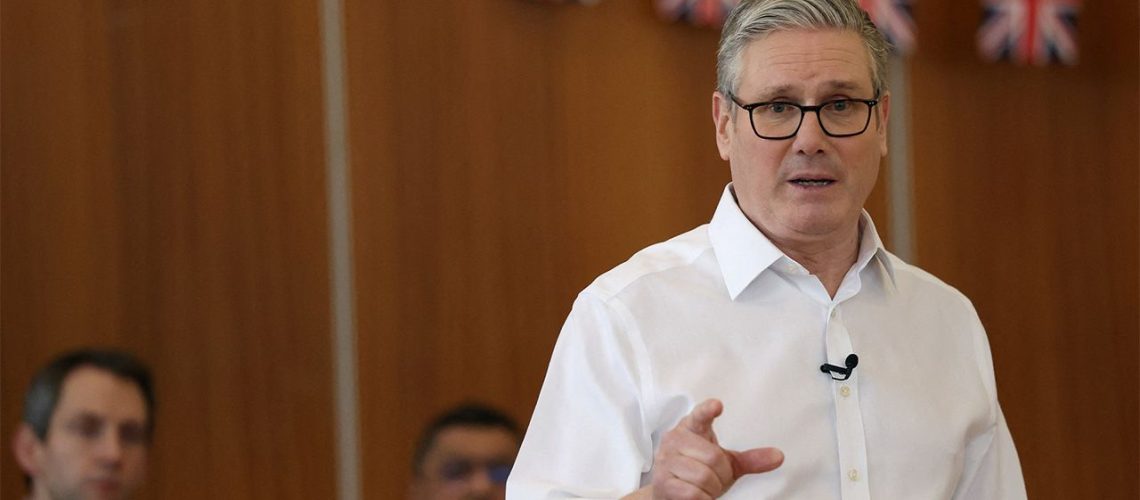 UK Prime Minister Keir Starmer delivers a speech in front of brown wall draped in Union Flags.