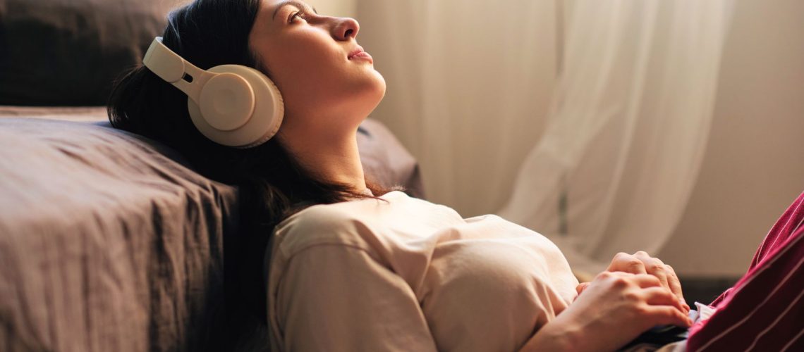 A young brunette woman relaxes against her bed while listening to a podcast through headphones.