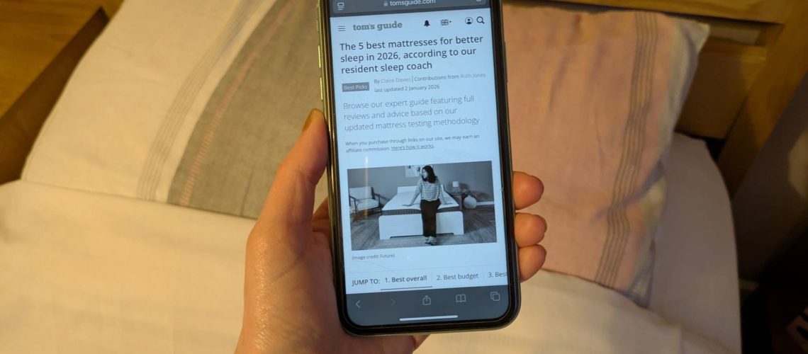 This image shows a hand holding an iPhone showing the Tom's Guide best mattress page in grayscale. In the background is a bed and pillows