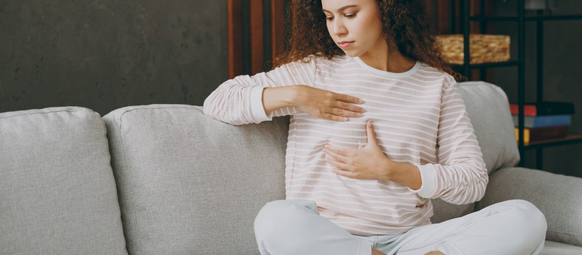 Full body young woman wearing casual clothes sits on grey sofa couch hold touch breast check health.