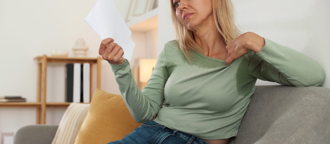 Woman waving paper sheet to cool herself during hot flash on sofa at home