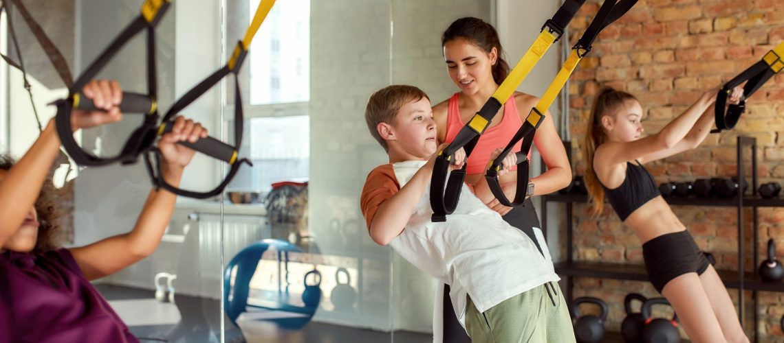 Portrait of female trainer helping teenage boy while he is training using fitness straps in gym