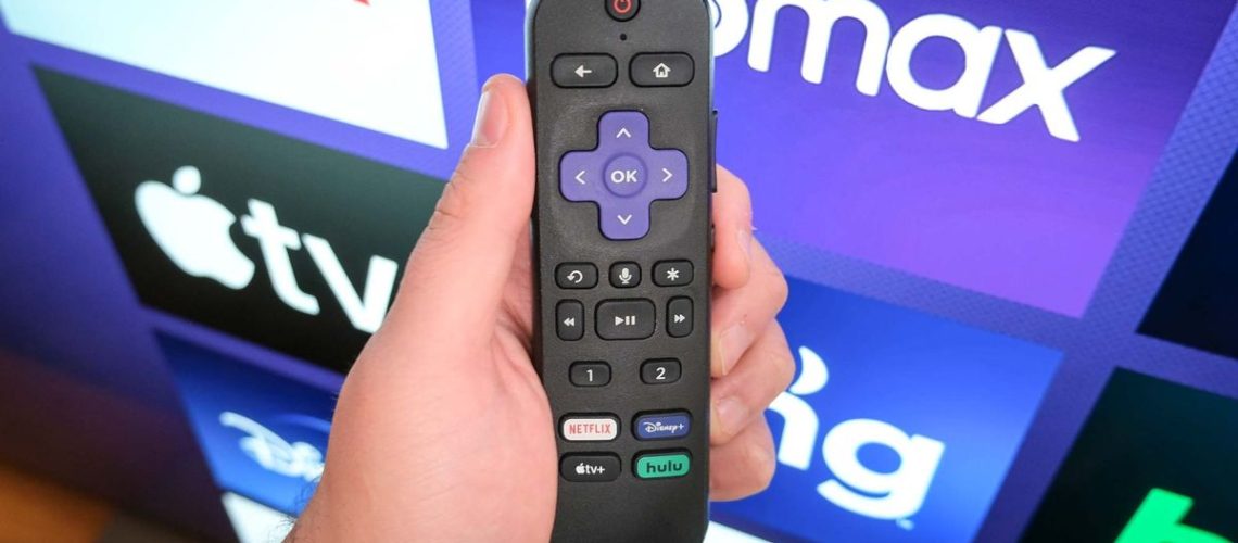 A Roku remote in hand in front of a TV with the Roku home page.