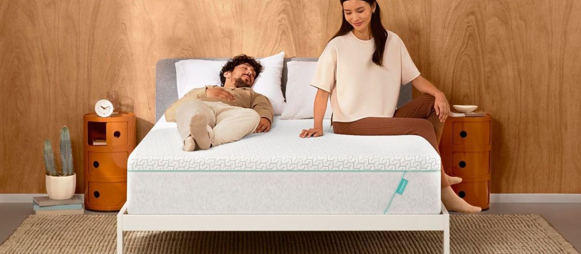 A couple sit on top of the Tuft &amp; Needle Original Hybrid mattress, which is placed on a white bed frame in a neutrally decorated bedroom