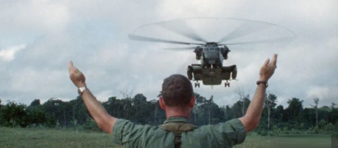 A man stands with his arms to the air as a helicopter approaches Jonestown