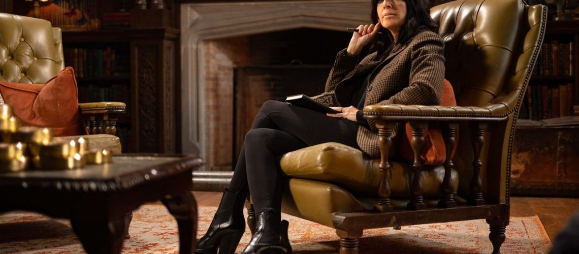 Host Claudia Winkleman sits by a fireside in a leatherbound chair in The Traitors U.K. season 2