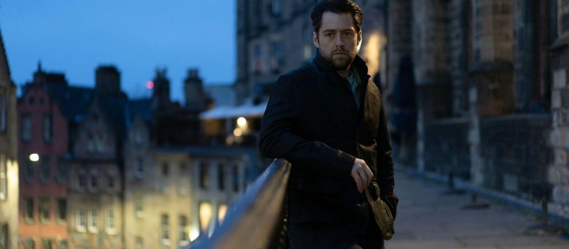 Detective Sergeant John Rebus (played by Richard Rankin) wearing a black jacket in Edinburgh old town