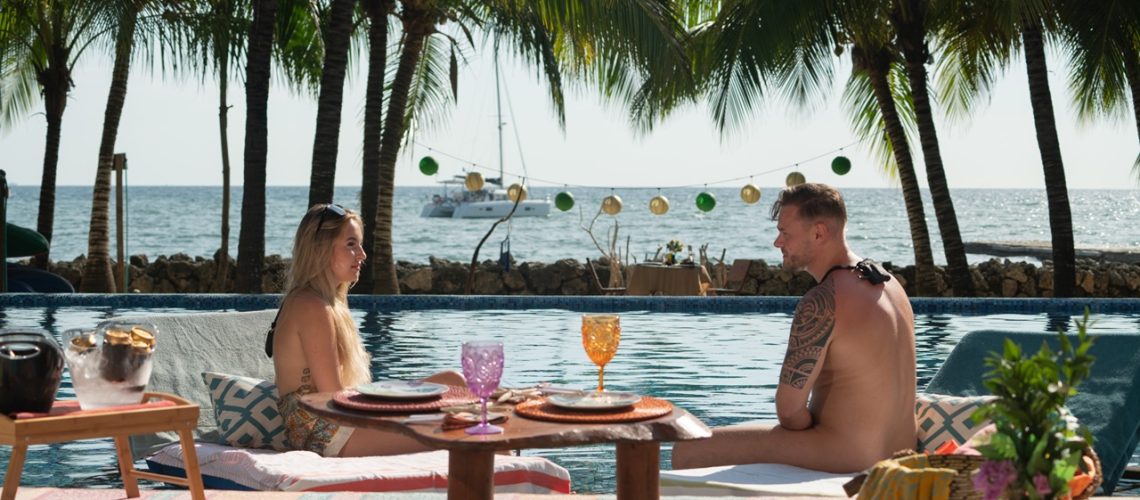 A naked woman and man sit chatting by the pool in a tropical resort in "Dating Naked Germany" season 2.