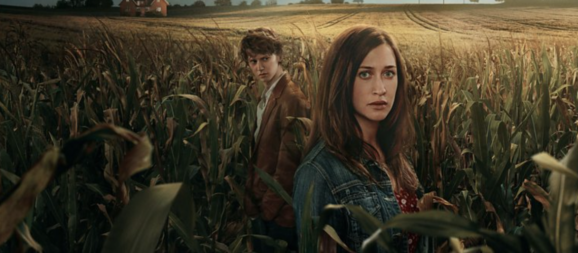 Vera (Julia Ragnarsson) and Isak (Erik Enge) stand in a wheat field with a house and wind turbines in the background in End of Summer