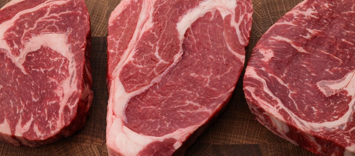 Close up three raw marbled ribeye beef steaks over end grain cutting board of wooden butcher block, high angle view, directly above.