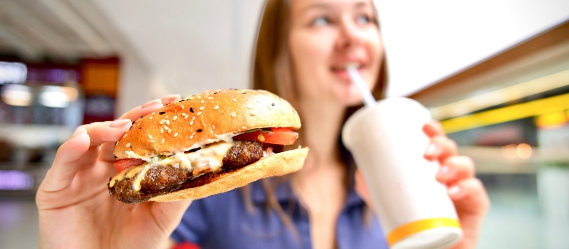 young woman holding big burger and cup of soda in shopping mall on food court.
