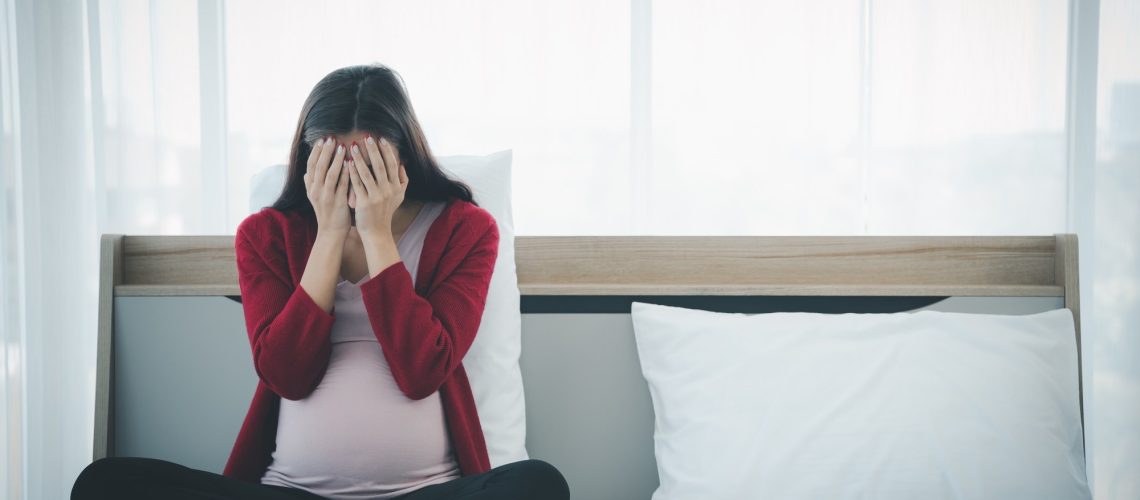 Pregnant woman in casual home outfit sitting on bed and feeling upset and sad
