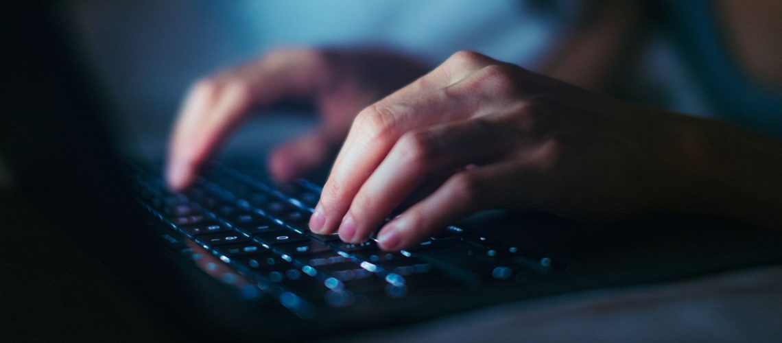 A close-up photograph of a person's hands typing on a backlit laptop keyboard