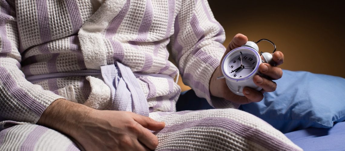 A man in a bathrobe holds an alarm clock in his hand