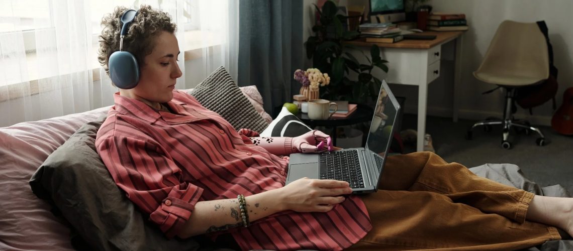 Caucasian young adult woman lying on bed using laptop and wearing headphones, holding smartphone in hand, surrounded by snacks, demonstrating signs of gadget addiction in bedroom