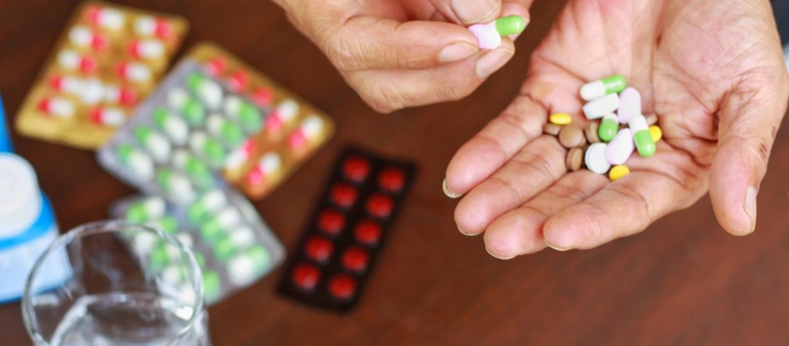Elderly man hands holding many pills(tablet and capsule). Packets of mediation and glass of water