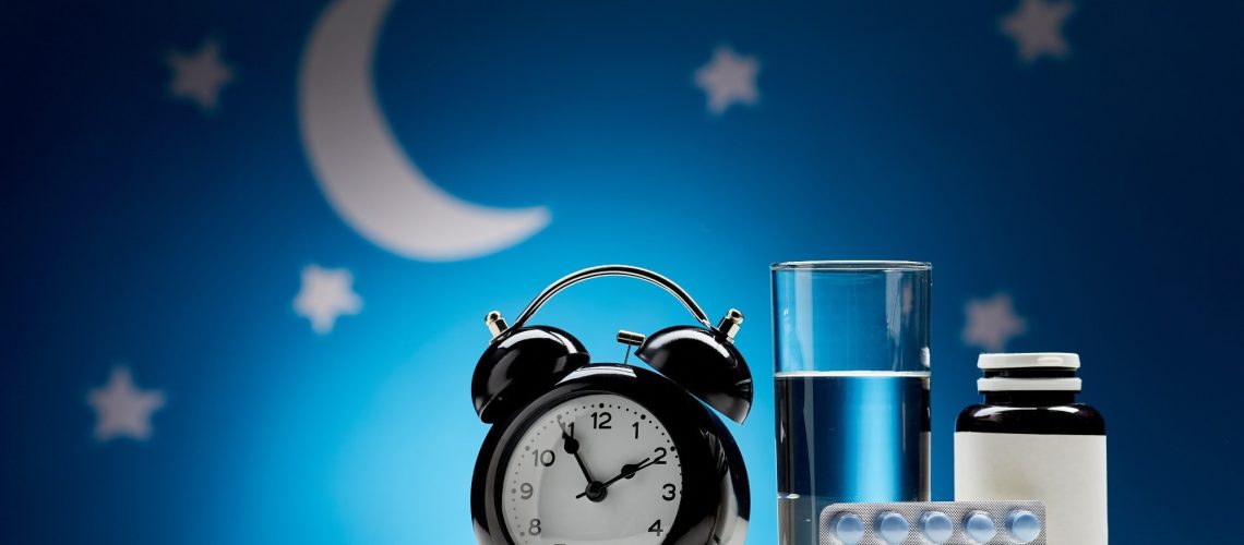 close up of alarm clock, glass of water, earplugs and sleeping pills over moon and night stars on blue background