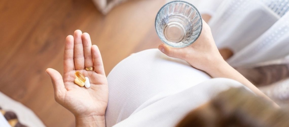 A pregnant woman in a white top holds a glass of water in one hand and a few prenatal vitamins in the other. The image is taken from a top-down perspective, highlighting her baby bump and the supplements she is about to take.