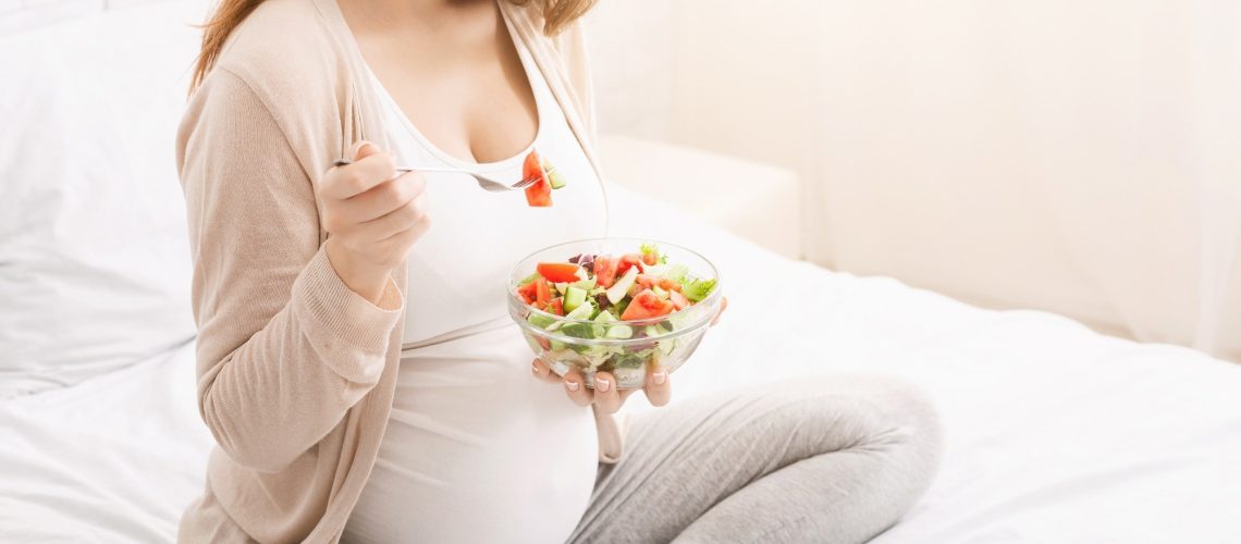 A pregnant woman is sitting on a bed, holding a bowl of salad in her hands