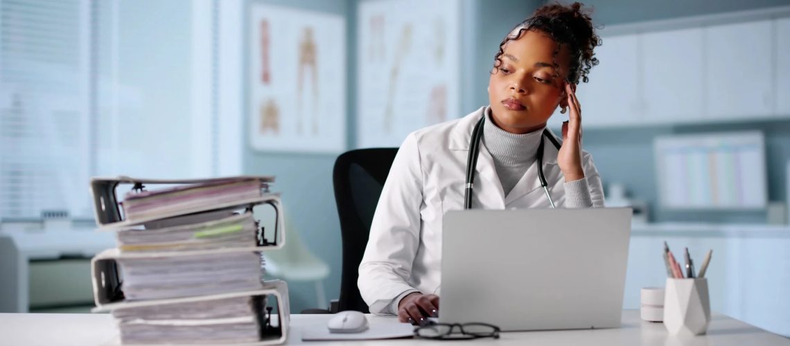 Exhausted Hospital Doctor Surrounded By Mountain Of Paperwork And Calculator.