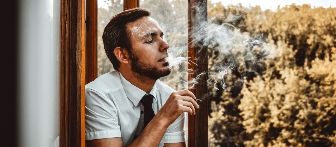 A man smokes a cigarette on the balcony against the background of autumn trees with golden foliage.
