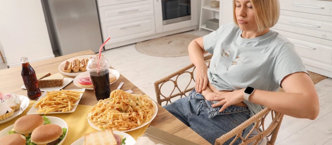 Woman sat at a dining table with lots of food on. Food on her shirt and her jeans are unfastened to show she has overeaten
