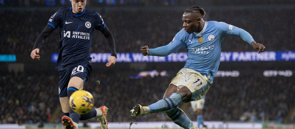 Cole Palmer (L), in navy blue kit, attempts to block a ball in from Jeremy Doku, in sky blue shirt and white shorts, at the Man City vs Chelsea live stream.