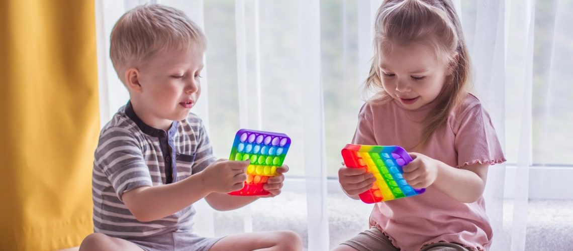 Blonde boy and girl children sit on the floor near the window and play silicone toy antistress pop it. Pop it sensory toy. Stress relief. Colorful anti-stress silicone sensors toy