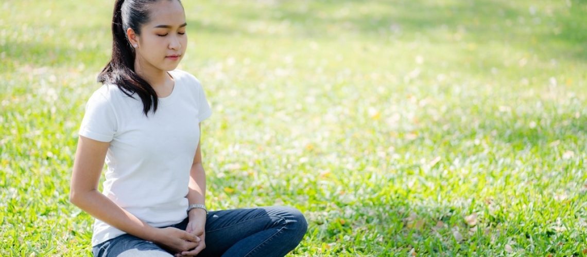 A young woman meditating peacefully outdoors on a sunny day, sitting cross-legged on green grass in a relaxed posture.