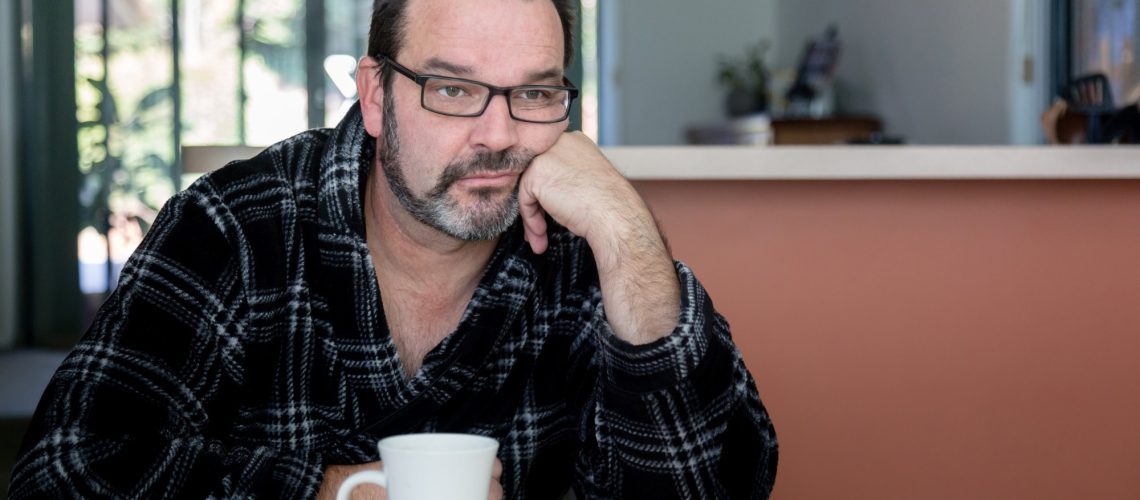 Middle aged man sitting at dining room table with coffee mug looking sad and depressed