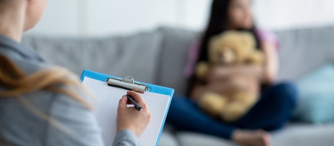 A blurred young girl sitting on a couch hugging a teddy bear, while a mental health professional in the foreground takes notes on a clipboard. The setting appears to be a therapy session in a well-lit room.