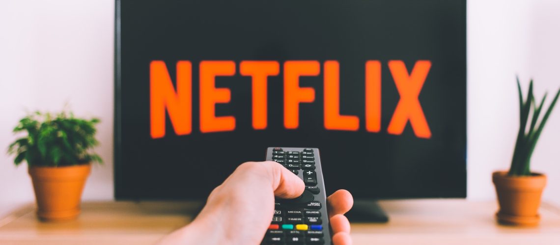 A TV with the Netflix logo sits behind a hand holding a remote
