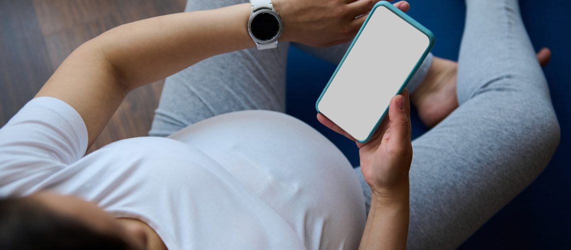 Top view pregnant woman checks smart watch to control pulse and heart rate, relaxing with smartphone