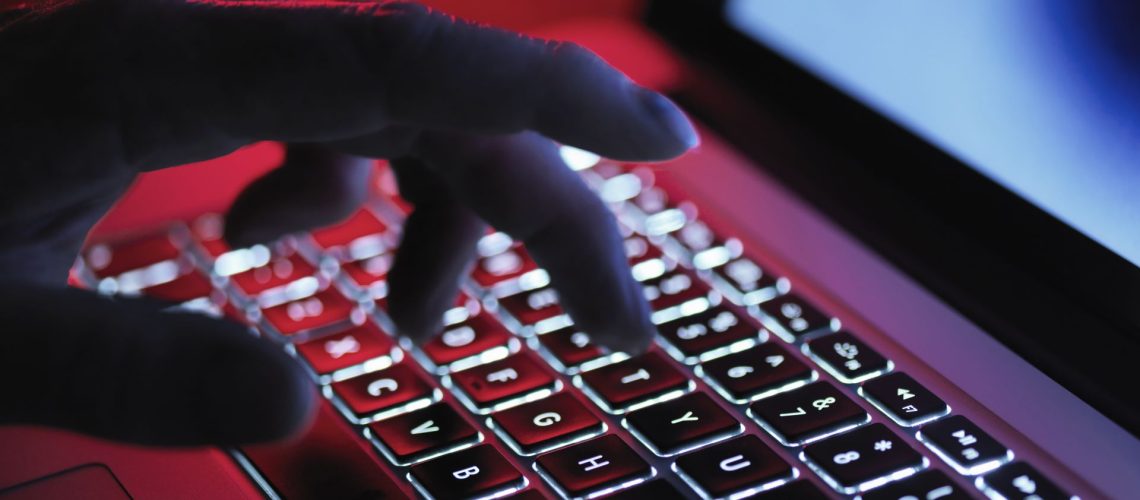A hand typing at a computer in a dark room, lit up by the laptop's keyboard LEDs and red LED light