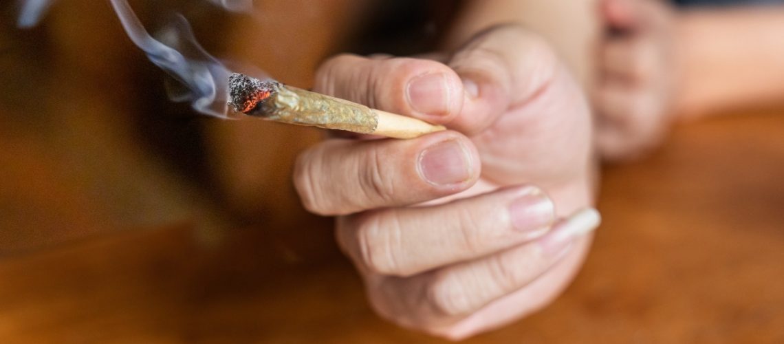 Finger holding lighted cannabis cigarette with smoke in dark room.