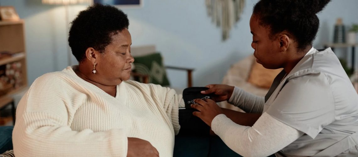 Female nurse wrapping cuff of automatic tonometer on elderly Black womans arm measuring blood pressure during health checkup visit at home