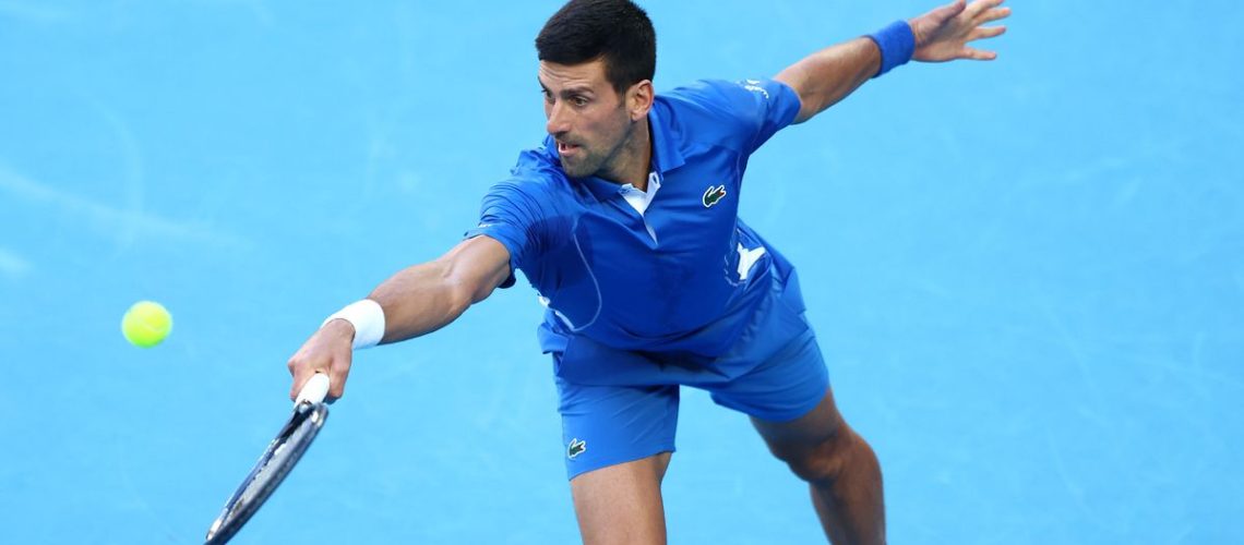 Novak Djokovic of Serbia, wearing royal blue tennis outfit, plays a lunging backhand on a blue court just prior to the Australian Open 2024