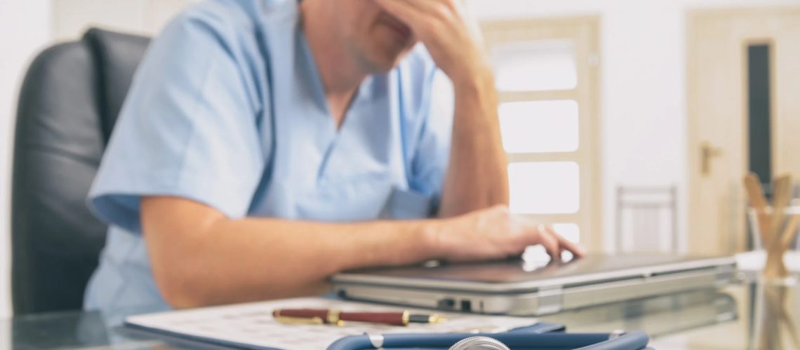 Overworked doctor sitting in his office with head in his hands. Focus on stethoscope in foreground