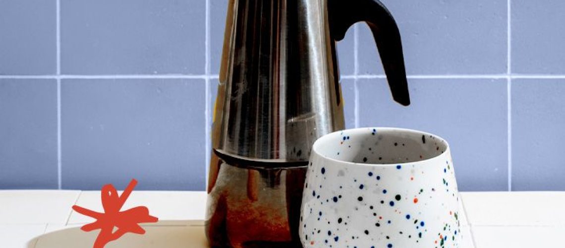 metal coffee pitcher and white cup with colorful speckles against a blue tile background