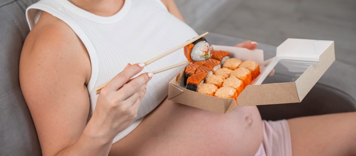 A pregnant woman sits on the sofa and eats rolls from a box
