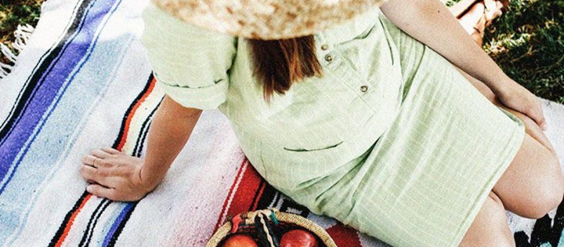 A pregnant woman sits on a blanket next to baskets of apples, nuts, and greens
